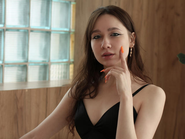 AdrianeWalter with long brown hair, black bra, orange nails, and gold earrings, pointing at her cheek with her index finger. She's looking at the camera with a slight smile.