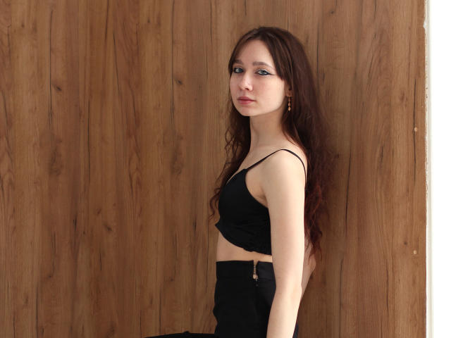 AdrianeWalter with long brown hair, wearing a black crop top and high-waisted black pants, standing against a wooden wall, looking to the side with a serious expression.