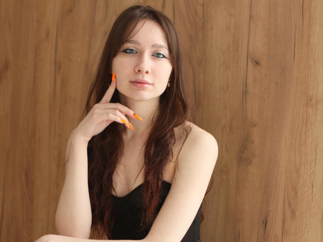 AdrianeWalter with long brown hair, blue eyes, and orange nails, touching her chin with a finger, wearing a black top, wooden background.