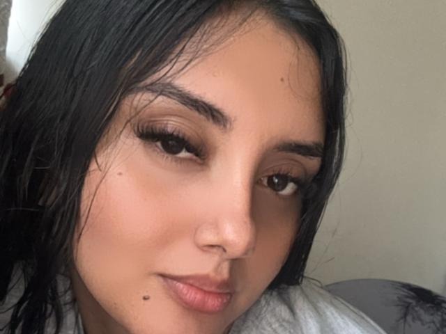 AmaliaDalton's face, dark wet hair, brown eyes with long eyelashes, subtle smile, light skin, small mole on right cheek, wearing gray top, looking at camera.