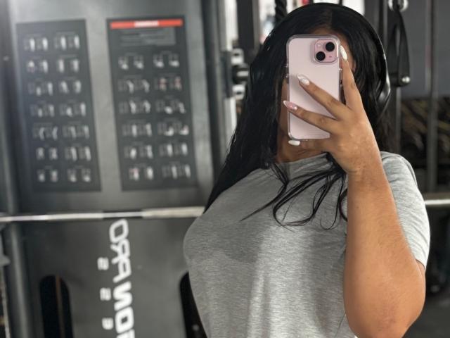 AmaliaDalton with wet black hair, holding pink phone, covering face, wearing grey shirt, showing big tits, standing in gym, reflecting in mirror, wearing headphones.