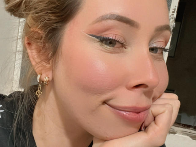 Il viso di JennyLawry, con un leggero blush rosa, un eyeliner nero intenso, occhi marroni e un sorriso appena accennato, con il mento appoggiato sulla mano, indossa orecchini dorati e ha i capelli biondi raccolti in uno chignon..