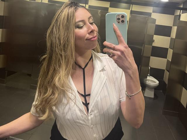 JennyLawry, con cabello rubio ondulado, vestida con una camisa blanca a rayas finas y un sujetador negro con tirantes, se toma un selfie frente a un espejo en un baño, mostrando sus pechos y una ligera sonrisa..