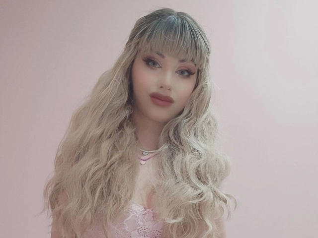 ChloeKiss-hot with long, wavy blonde hair, pink lipstick, and a cute pink lace top, looking sexy and confident. Her tits are small, and her ass is not visible.