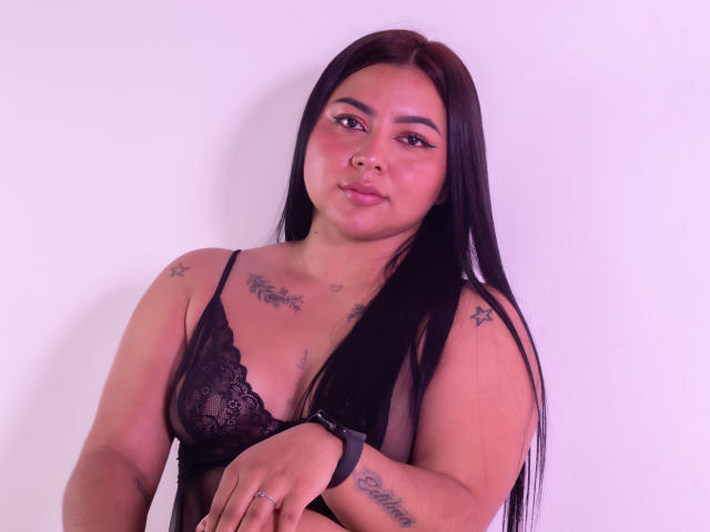 IvannaSmith69, con cabello negro largo, vestida con un sujetador de encaje negro, tiene tatuajes en el pecho y los brazos, un piercing en la nariz y un reloj, y mira a la cámara con una ligera sonrisa..