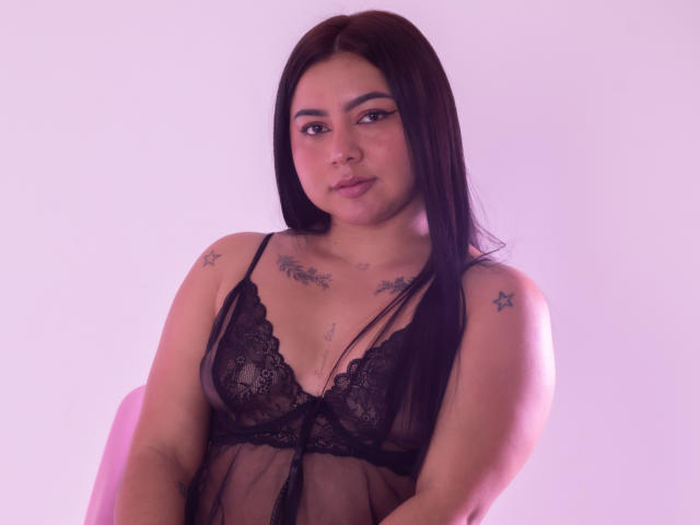 IvannaSmith69, con lunghi capelli neri, indossa un sottile vestitino nero in pizzo, che mette in mostra piccoli seni e tatuaggi sulle spalle e sul petto. Ha un'espressione neutra, con una luce rosa..