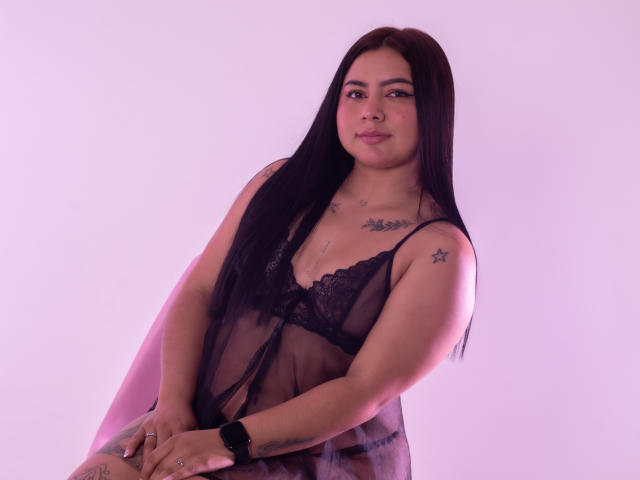 IvannaSmith69, con lunghi capelli neri, indossa un top di lingerie nero trasparente, mostrando piccoli tatuaggi sulla spalla e sul petto, seduta con un sorriso discreto e un orologio nero..