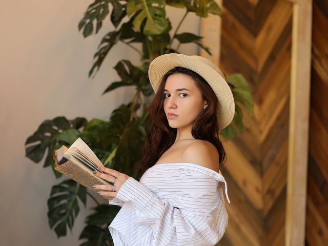 MeryMoris-hot with wavy brown hair, wearing a white striped shirt off-shoulder and a beige hat, holding a book, looking over her shoulder, standing by a green plant.
