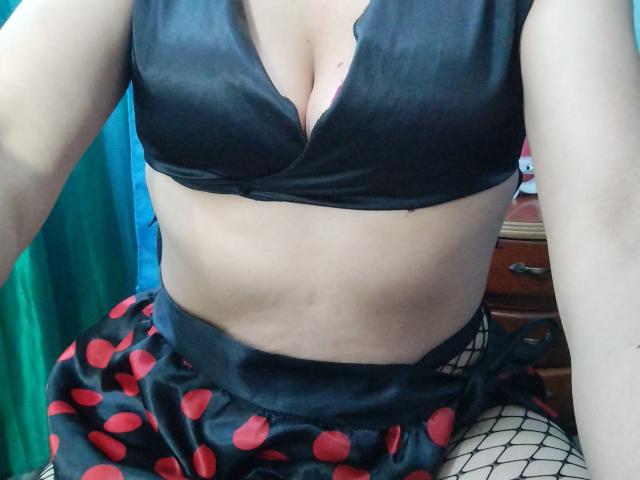 HanaCuteX wearing a black bra with deep cleavage, black and red polka dot skirt, and fishnet stockings, showing her bare midriff and slightly tan skin.