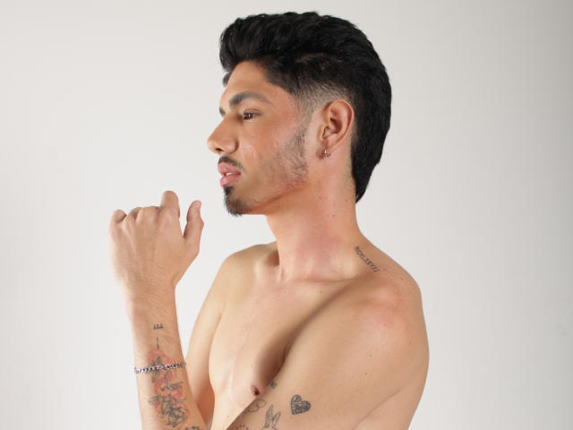 Shirtless FabrizzioHot with dark hair, small tattoos, ear piercing, and a bracelet, making a "rock on" hand gesture, showing a slight smile and visible nipples.