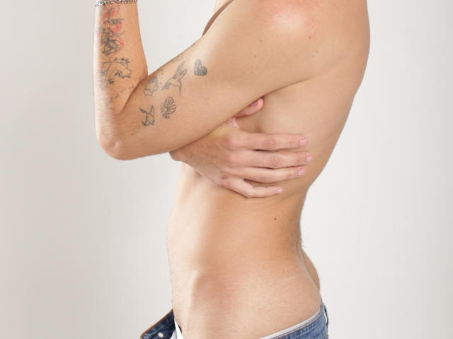 FabrizzioHot's shirtless, side profile with tattoos on his arm, hand covering his ass, wearing blue jeans, light skin, and a silver bracelet.