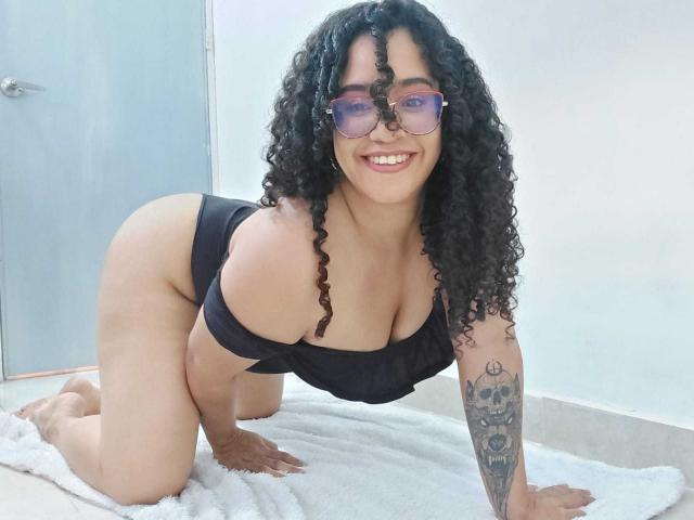 Kalith, een mollige vrouw met krullend zwart haar en een bril, staat op handen en voeten in een zwart bikini, lachend naar de camera, waarbij ze haar grote borsten en getatoeëerde arm laat zien..