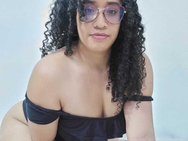 Kalith, con cabello negro rizado, gafas y una blusa negra con un hombro descubierto que muestra sus pechos, inclinada hacia adelante, sonriendo ligeramente y usando gafas de color rosa..