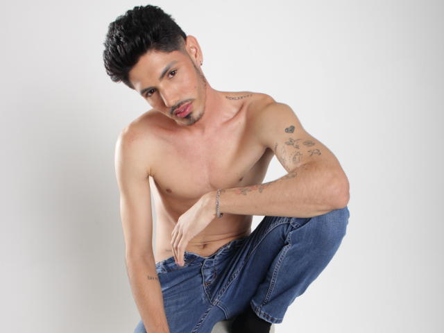 Shirtless FabrizzioHot with tattoos, dark curly hair, and a slight beard, crouching in blue jeans, looking down with a relaxed, slightly sexy expression.
