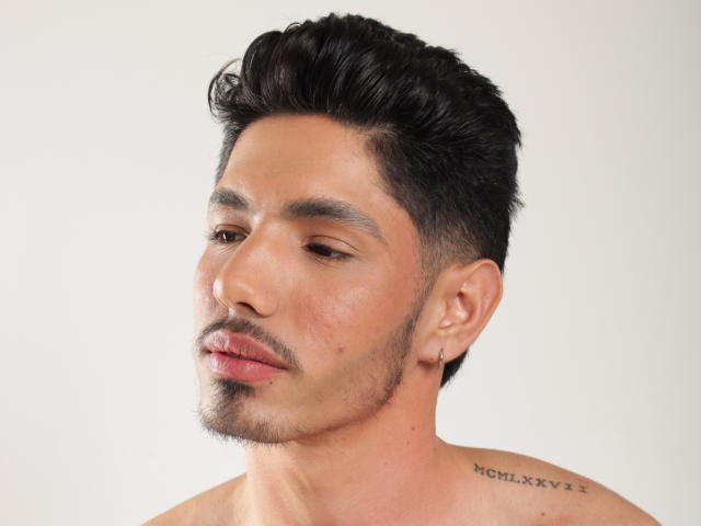 FabrizzioHot with short, dark, styled hair, slight stubble, and a small goatee. He's shirtless, has a tattoo on his right shoulder, and a small earring.