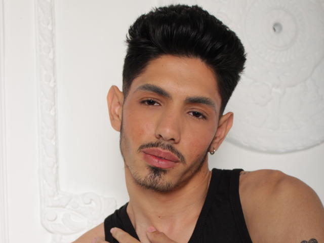 FabrizzioHot has a medium skin tone, short black hair, a small goatee, and a black tank top. He's looking at the camera with a slight pout, wearing a tiny earring.