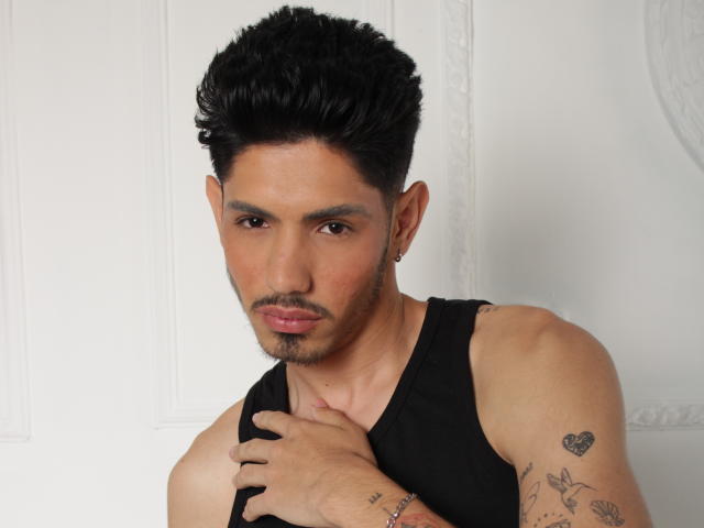 FabrizzioHot with short, curly black hair, tan skin, and a black tank top, holding his chest with tattoos on his right arm. He's looking serious, with a small earring.