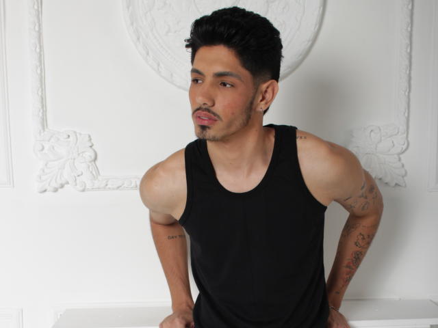 FabrizzioHot with short black hair, light brown skin, tattoos on both arms, and a black tank top, standing with hands on white surface, looking to the side.