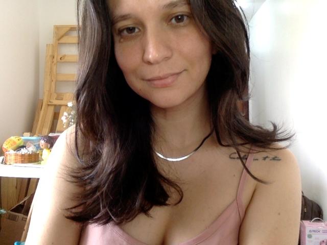 SweetBea with wavy dark brown hair, pink tank top, small tits, tattoo on right shoulder, smiling, white walls, wooden ladder in background, black necklace, soft light.