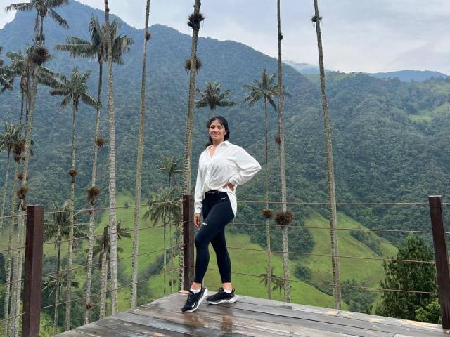 MaddieDemie with ass in black pants, white shirt, standing on wooden platform, palm trees in background, wearing black and white sneakers, hands on hips, looking at camera.