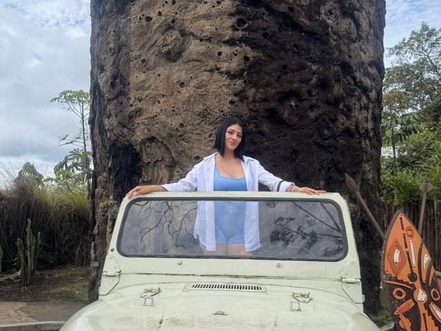 MaddieDemie with medium tits, wearing a blue bikini and white shirt, leaning on a white, old jeep against a rocky wall, black shoulder-length hair, standing in a forest.
