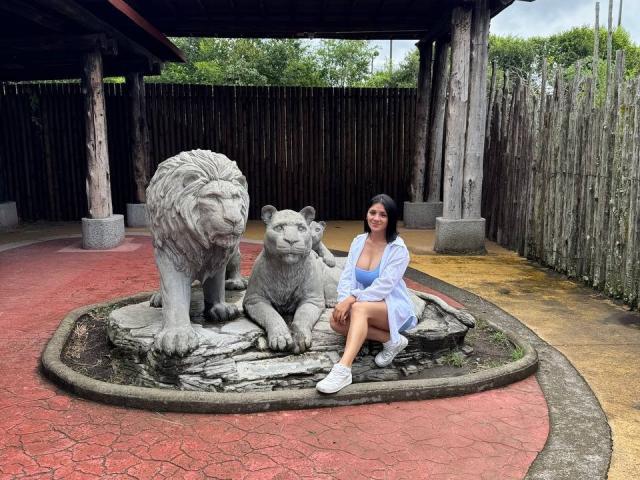 MaddieDemie with tits visible, wearing a white shirt and blue dress, sitting next to a stone lion and lioness sculpture in a circular red and yellow paved area.