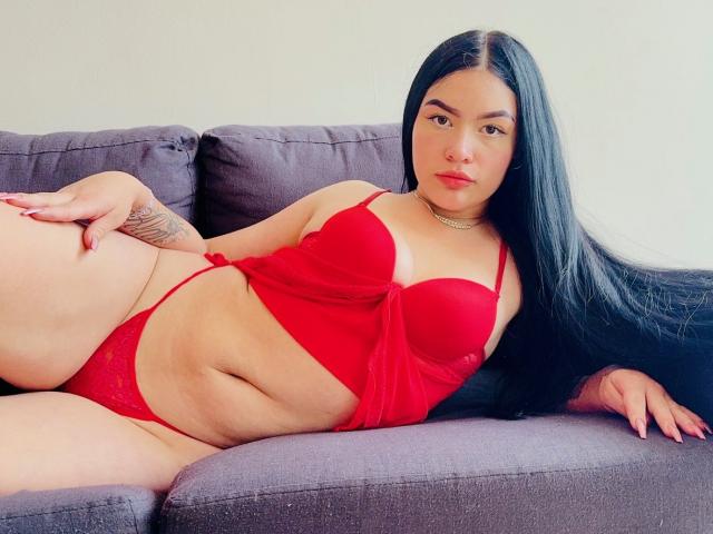 AdeleMontana, con cabello negro largo, lencería roja y un tatuaje en la mano derecha, acostada en un sofá gris, mostrando sus pechos y vulva, con una expresión neutra..