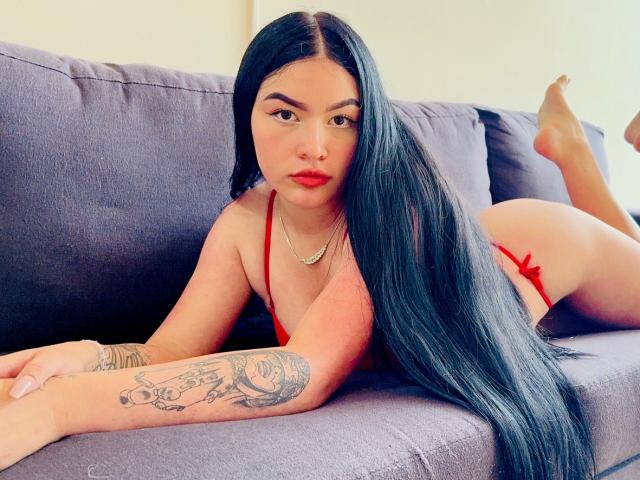 AdeleMontana with long black hair, red lingerie, red lipstick, and tattoo on her right arm, lying on a gray couch, looking at the camera, her ass raised.