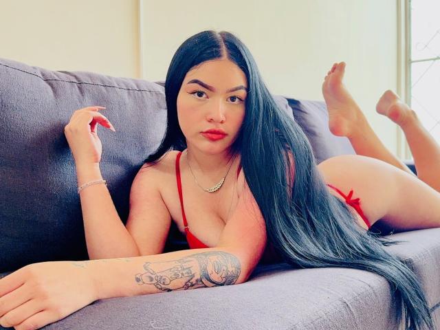 AdeleMontana, con lunghi capelli neri, biancheria intima rossa e un tatuaggio sul braccio destro, sdraiata su un divano grigio, con le gambe sollevate, che mostra il sedere, e che guarda verso la fotocamera..