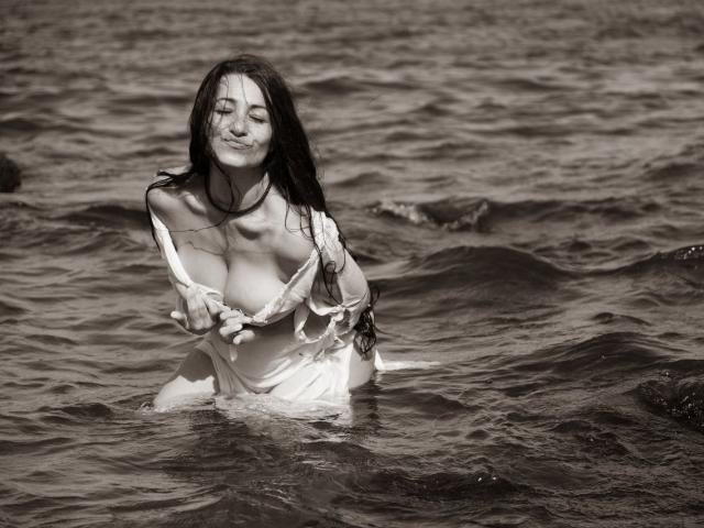 GinaONeon with wet long dark hair, smiling, holding down her wet white dress to expose her large tits, standing in a sunlit, rippling, dark water ocean.