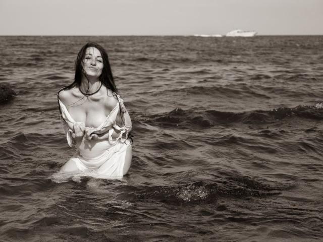 GinaONeon with long dark hair, smiling, standing waist-deep in water, wearing a white, partially wet, fringed top, exposing her tits, with a boat in the background.