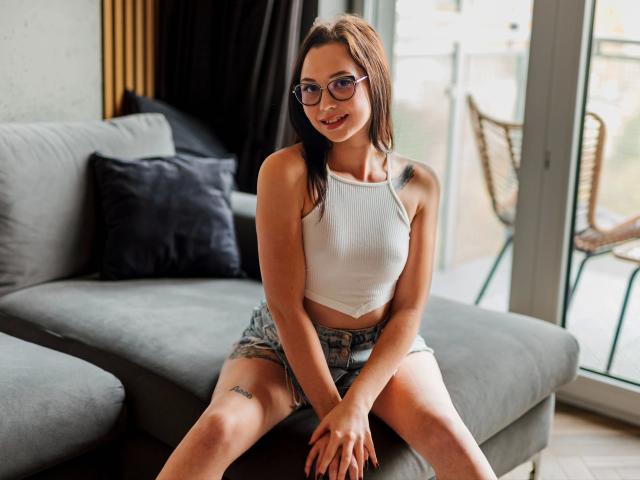 KlaraBest, con gafas, lleva un top blanco y pantalones cortos vaqueros, está sentada en un sofá gris, mostrando un tatuaje en su muslo derecho, y sonríe a la cámara..