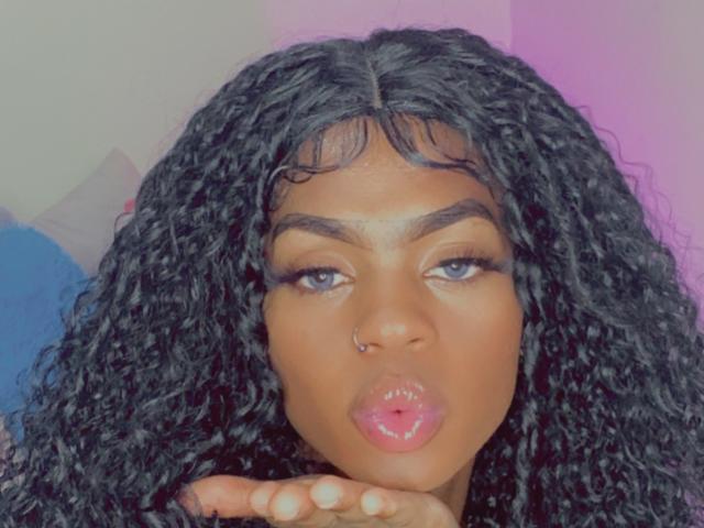 TayssaMaravilhia has curly black hair, a nose ring, and puffy pink lips, making a puckered face with her hand near her mouth. Her eyes are half-open.