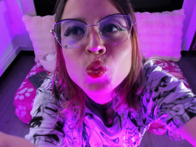 MelaniBecker with pouty red lips, wearing glasses, and a floral shirt, has pink and purple lit hair, sitting on a pink pillow, looking at the camera.