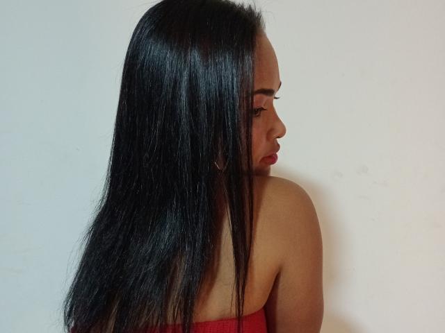KatherinCatis a de longs cheveux noirs et brillants, porte un haut rouge sans bretelles et du rouge à lèvres rouge, et regarde sur le côté, montrant ses fesses. Elle porte de petites boucles d'oreilles dorées..
