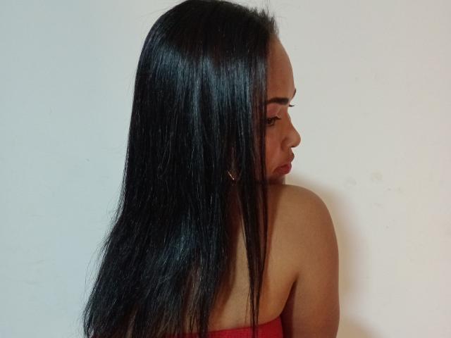 Katherin tem cabelos longos e lisos, pretos, um top vermelho sem alças, brincos dourados e uma expressão séria, olhando para a direita. Suas nádegas estão parcialmente visíveis..