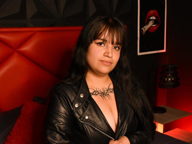 MarianeGrey with black leather jacket, low cleavage, tattoo on chest, long black hair, bangs, slight smile, sitting on red bed, red pillow, red and black decor.