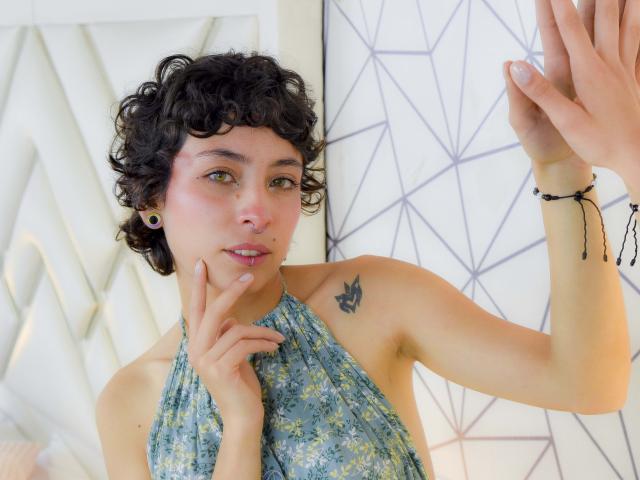EmilyAllen with curly black hair, wearing a floral halter top, has small tits and a butterfly tattoo on her right shoulder, raising her hands, and biting her finger.