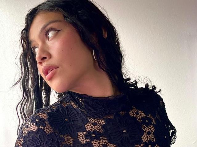 Zamantta has wavy black hair, wearing a black lace top with floral patterns, looking to the side with pouty lips, wearing subtle winged eyeliner, and hoop earrings.