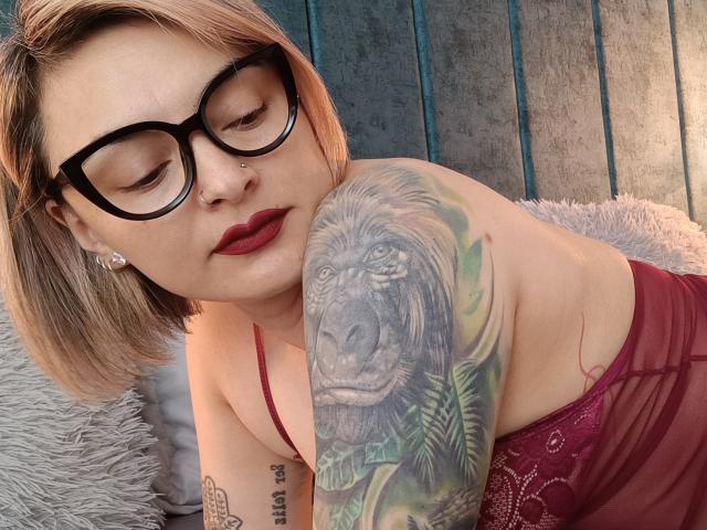 KatParadise, avec des lunettes, des cheveux blonds courts, du rouge à lèvres rouge et un haut en dentelle rouge, appuyée sur un coussin gris et moelleux, montrant un tatouage détaillé d'un singe réaliste sur son bras..