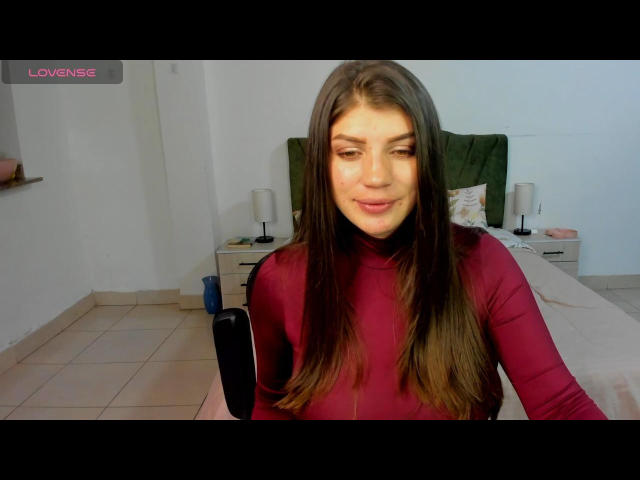 AnneCamile with long brown hair, wearing a red turtleneck, smiling at the camera, her tits slightly visible under the tight top, in a tiled room with a bed.