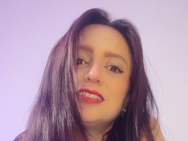 CatalinaMonyque a les cheveux noirs et lisses, porte du rouge à lèvres rouge, a les yeux marron, regarde légèrement sur le côté avec un sourire discret, et porte de petites boucles d'oreilles..