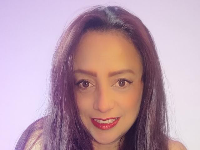 CatalinaMonyque a les cheveux brun foncé de longueur moyenne, porte du rouge à lèvres et sourit avec ses yeux marron. Ses seins sont légèrement visibles sous un haut sans manches..