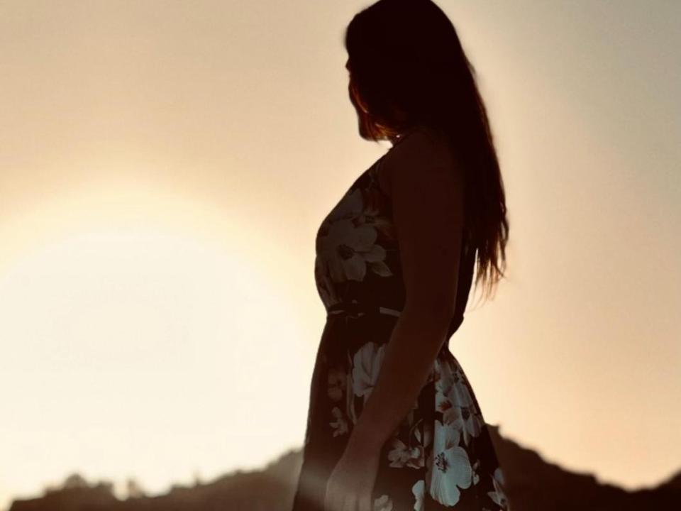 Silhouetted OctaPussy with long brown hair, wearing a floral dress, standing in profile against a sunset, her ass slightly curved, backlit, glowing softly.