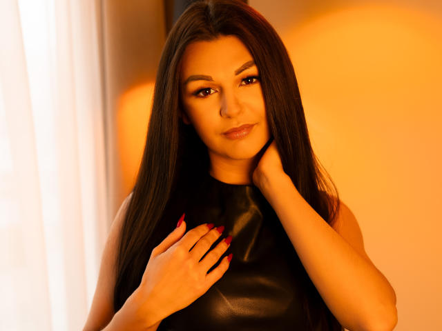 DariaHypnotic with long dark hair, wearing a black leather sleeveless top, red nails, hand on neck, warm orange light, smiling subtly, looking at camera.