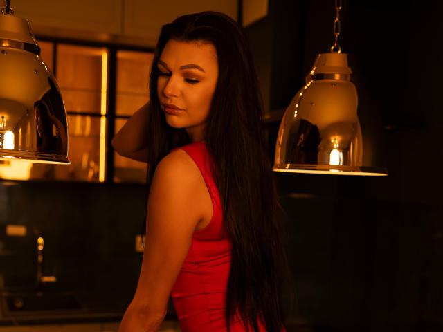Daria, con su largo cabello negro y un vestido rojo, mira por encima del hombro, de pie en una cocina tenuemente iluminada con dos lámparas colgantes. Su trasero es ligeramente visible..