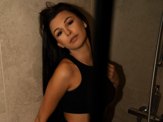 Daria, met lang zwart haar, draagt een zwarte crop top, waarbij haar borsten zichtbaar zijn, en staat tegen een donkere muur, kijkend naar de camera met een lichte glimlach..