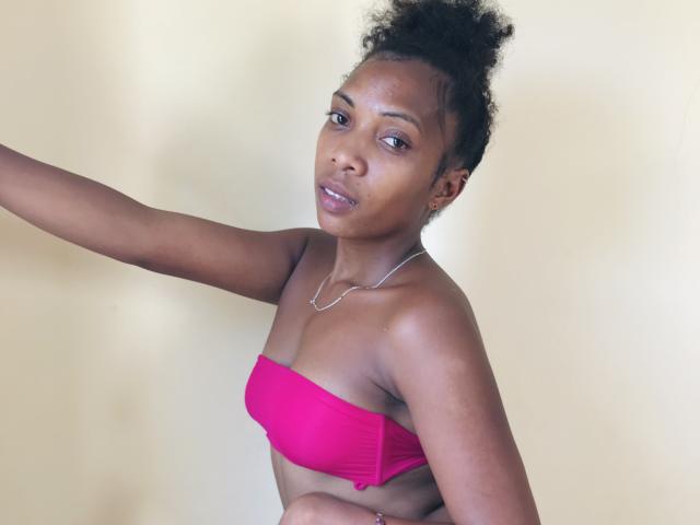 AntonniaMagik with dark brown skin, curly black hair in a bun, wearing a pink strapless top, silver necklace, and a subtle smile, arm extended to the side.