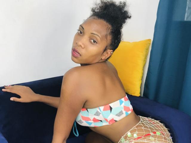 AntonniaMagik with curly hair, brown skin, and pink lipstick, wearing a geometric-patterned tube top and fishnet shorts, looking over her shoulder, sitting on a blue chair.