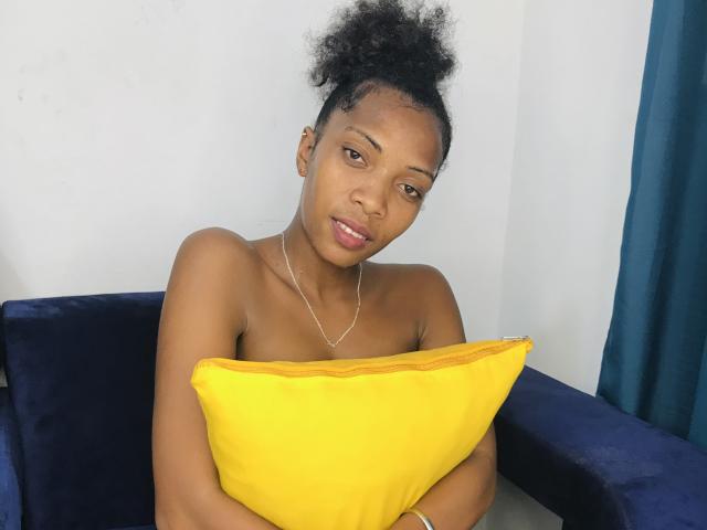 AntonniaMagik with dark skin, curly black hair in a bun, holding a yellow pillow, sitting on a dark blue chair, wearing a silver necklace, looking at the camera.